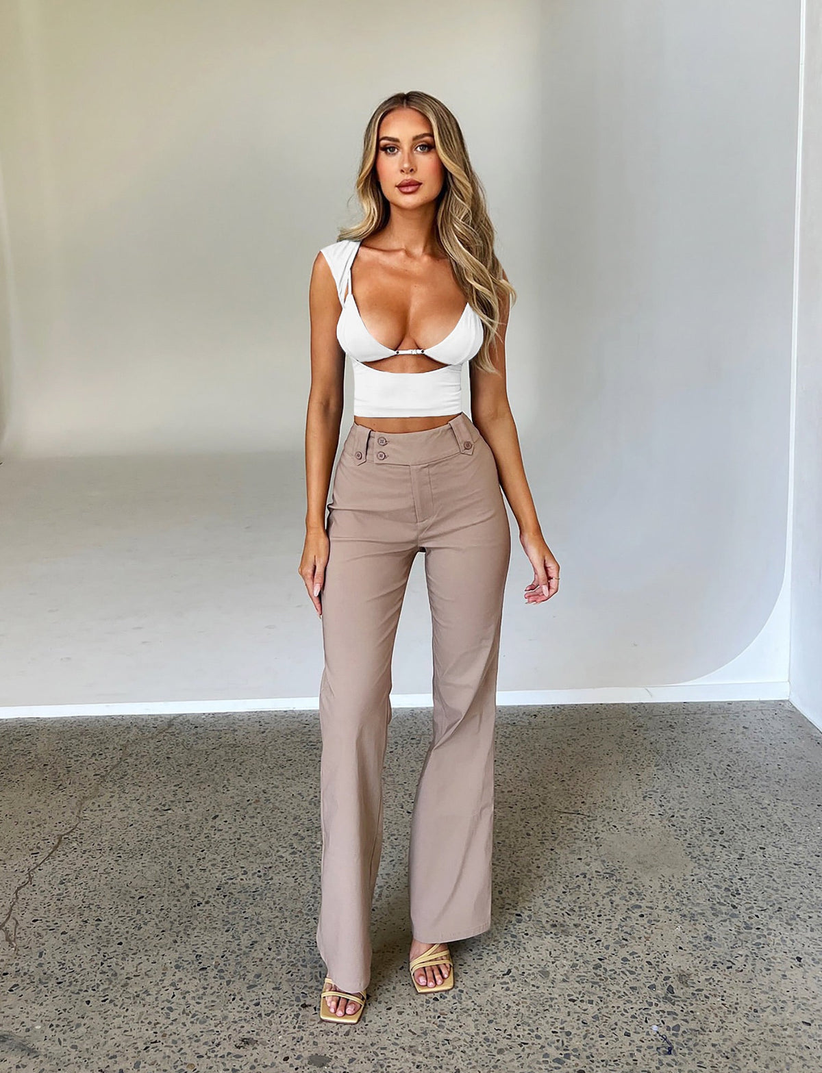 A woman with long hair stands posing against a plain backdrop. She is wearing a white, fitted crop top with cutouts and the KITTIE HIGH RISE PANT - BEIGE : TAN from TIGERMIST. She is also wearing beige open-toe sandals. Her expression is neutral, and her hands are relaxed by her sides.