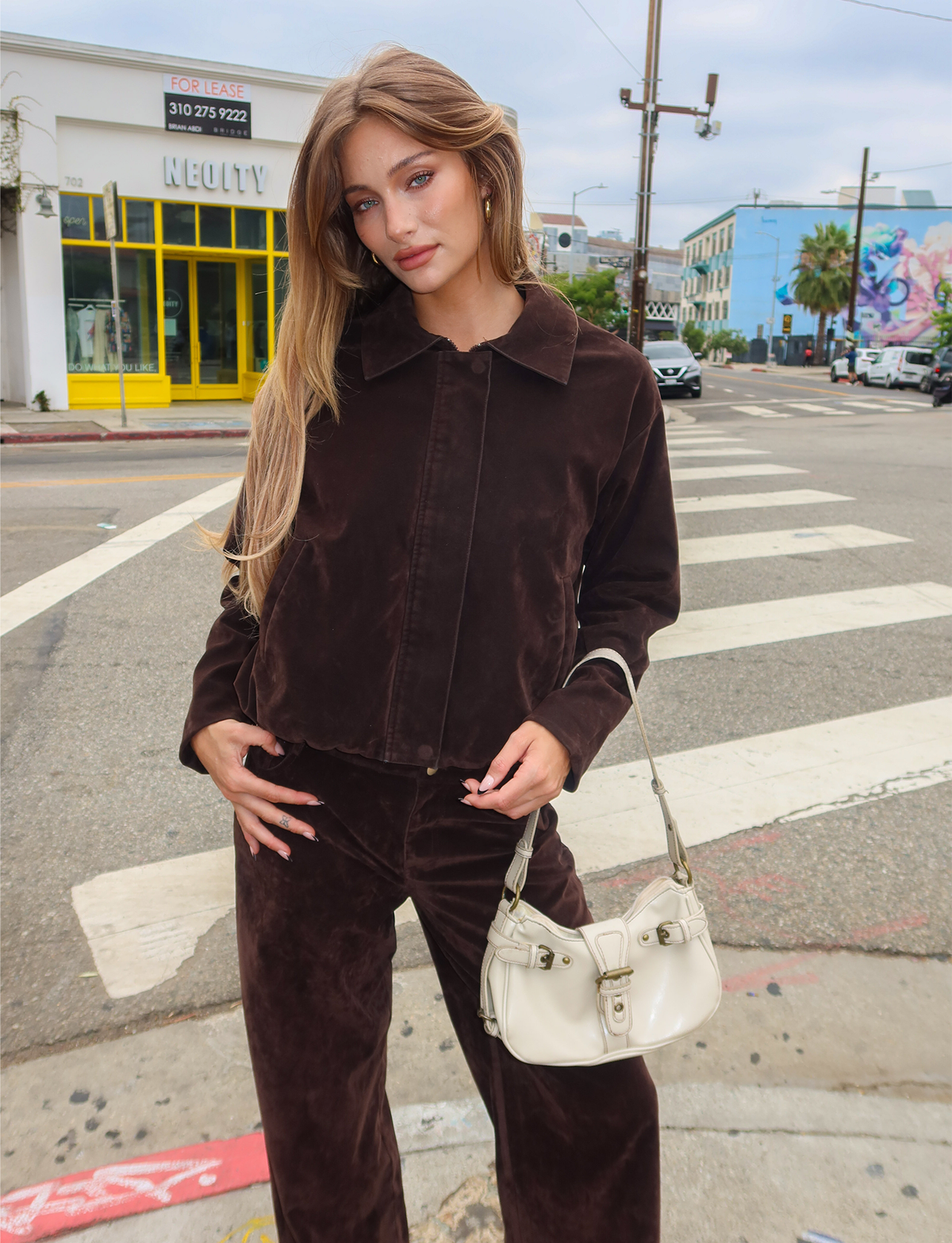 A woman with long hair stands at a city crosswalk holding a white handbag, wearing TIGERMIST’s CAMELLIA BOMBER JACKET in chocolate suede with matching pants. Colorful buildings and street art are visible in the background.