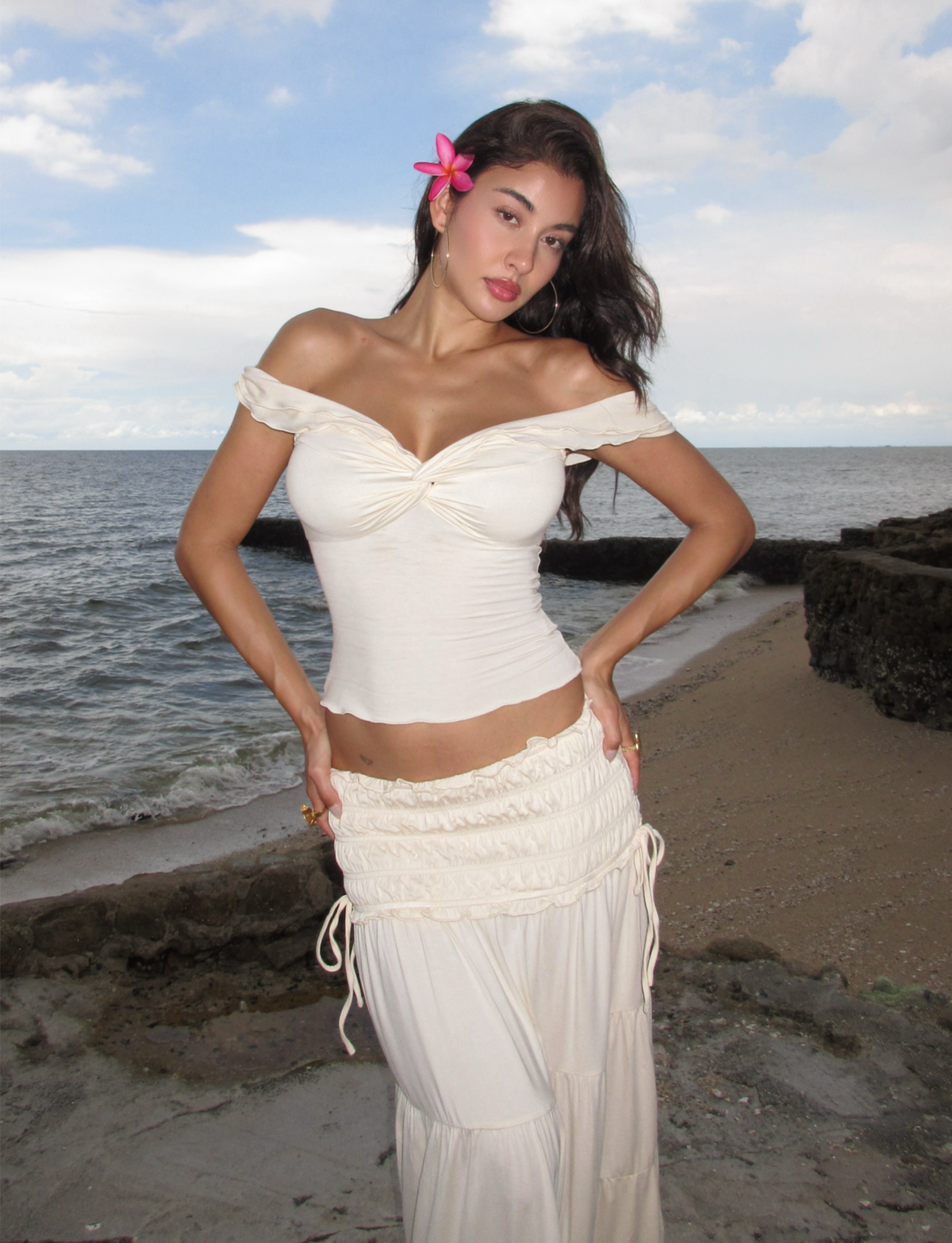A woman stands on a beach wearing the TIGERMIST DENALI TOP in Bone, an off-the-shoulder neutral top paired with a matching long skirt. A pink flower adorns her hair as the ocean and cloudy sky form the backdrop.
