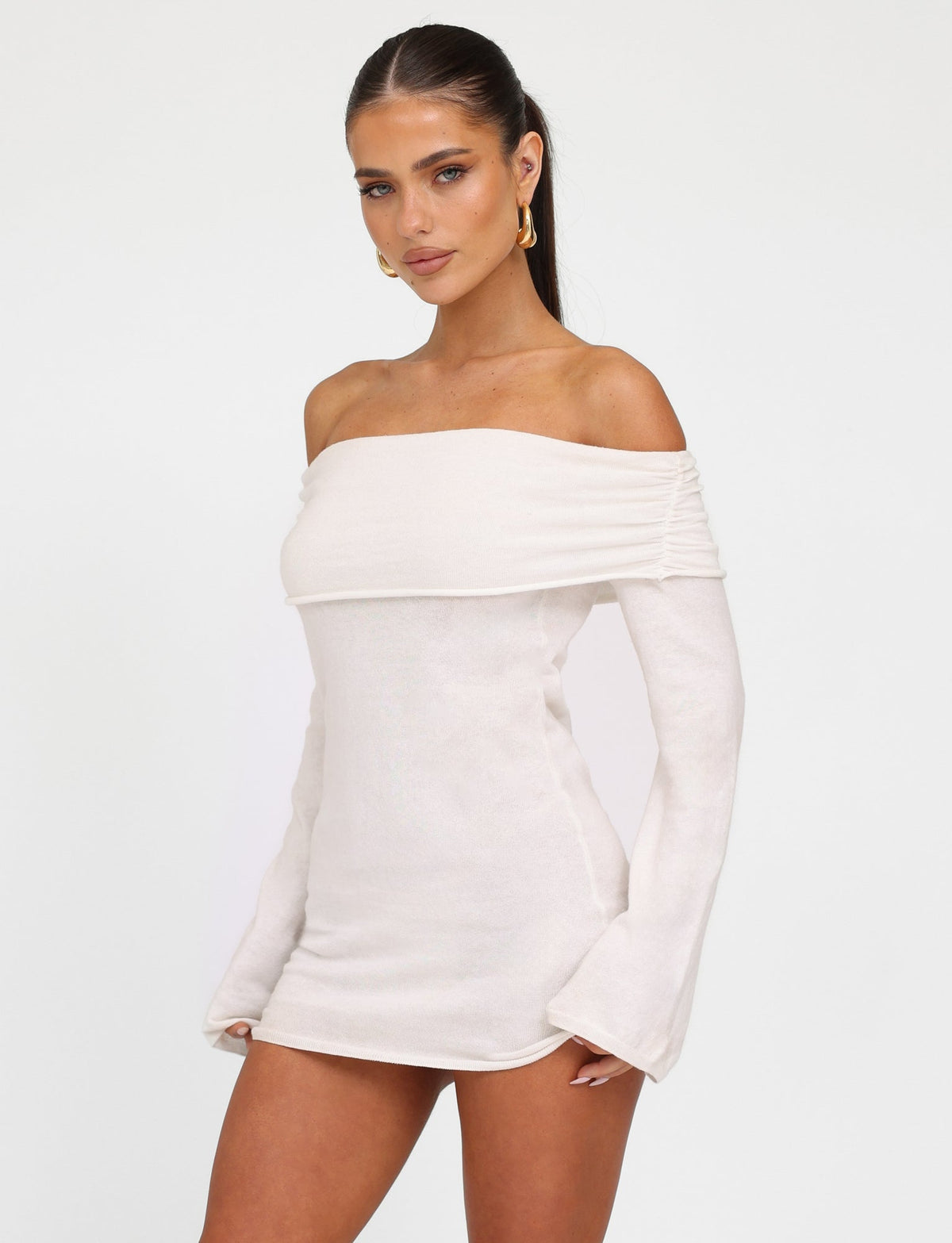 A woman with long dark hair is wearing the DIEDRE JUMPER DRESS - WHITE : CREAM from TIGERMIST. She stands confidently with her hand on her hip, looking directly at the camera. She accessorizes with large gold hoop earrings against a plain white background.