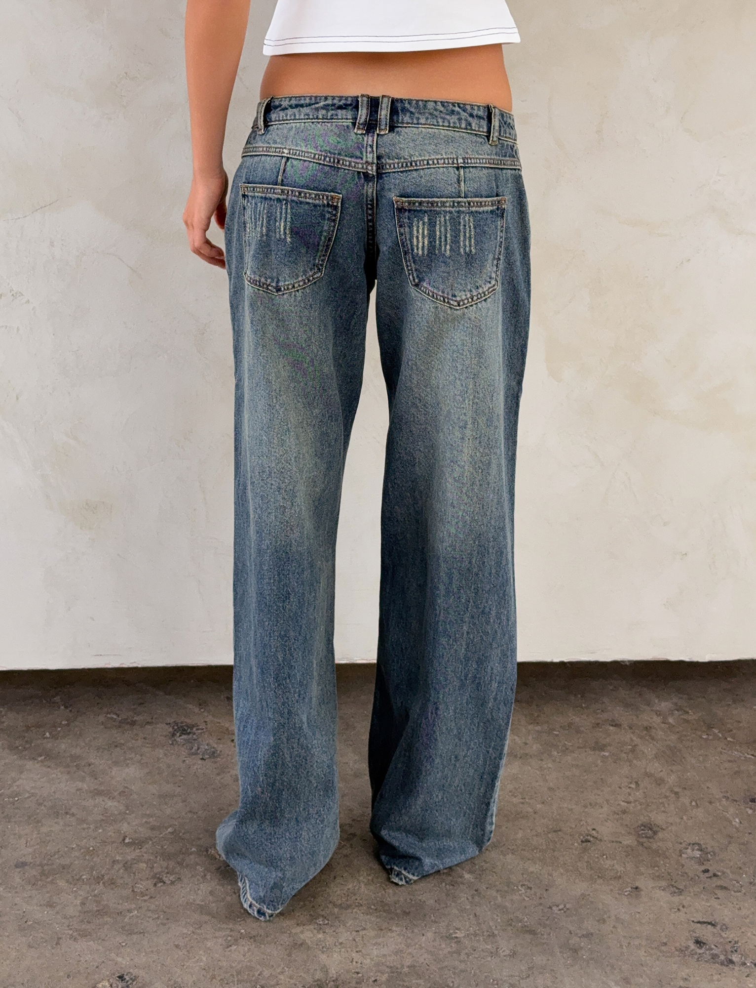 A person wearing the I.AM.GIA DYLAN JEAN in blue denim and a white top stands with their back to the camera against a light-colored textured wall and concrete floor.