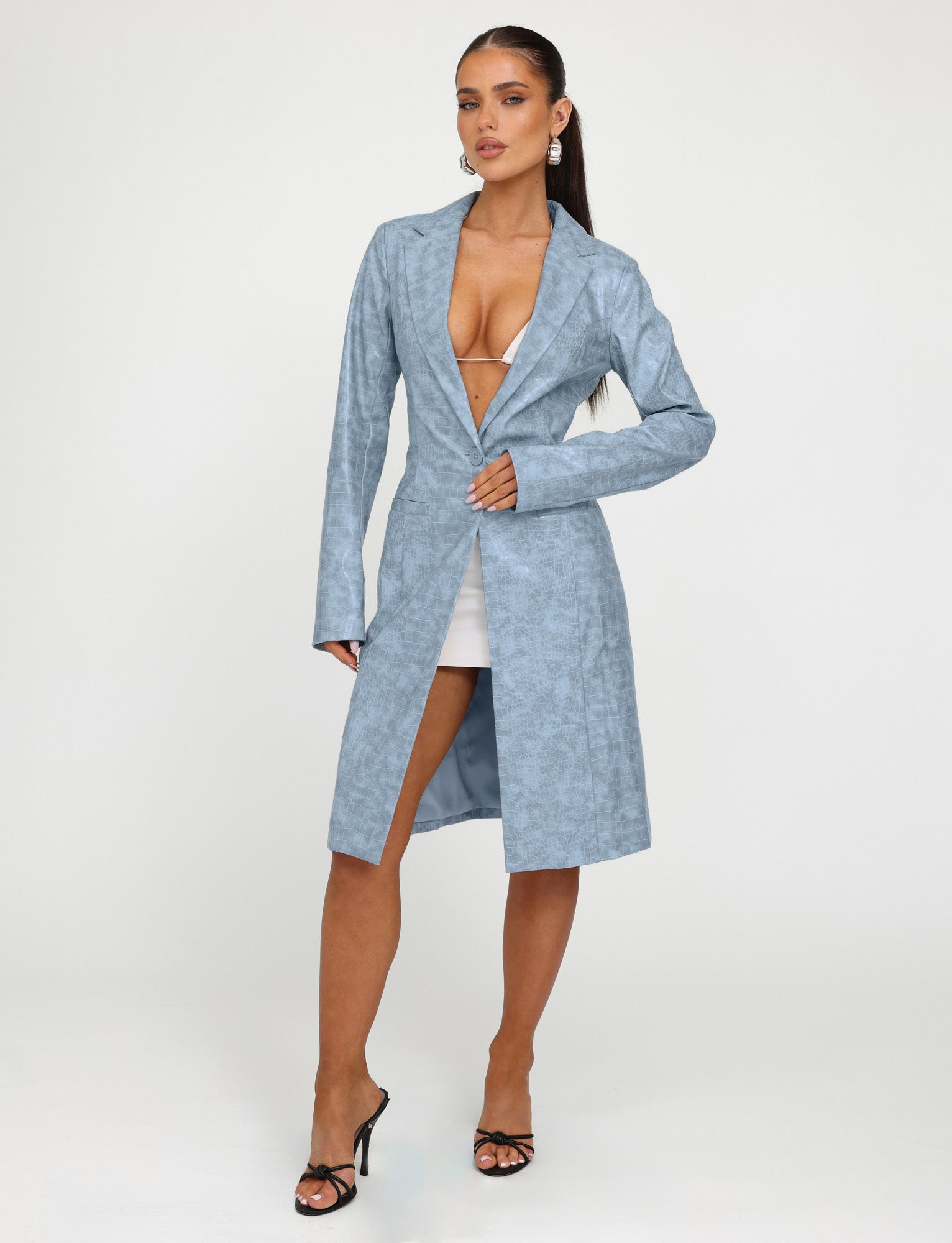 A woman stands against a plain, off-white background, wearing the TigerMist EMERSYN TRENCH COAT - BLUE : DUSTY BLUE with a deep V-neck over a white mini skirt. She accessorizes with dangly earrings and black high-heeled sandals. She gazes confidently at the camera with one hand on her coat.
