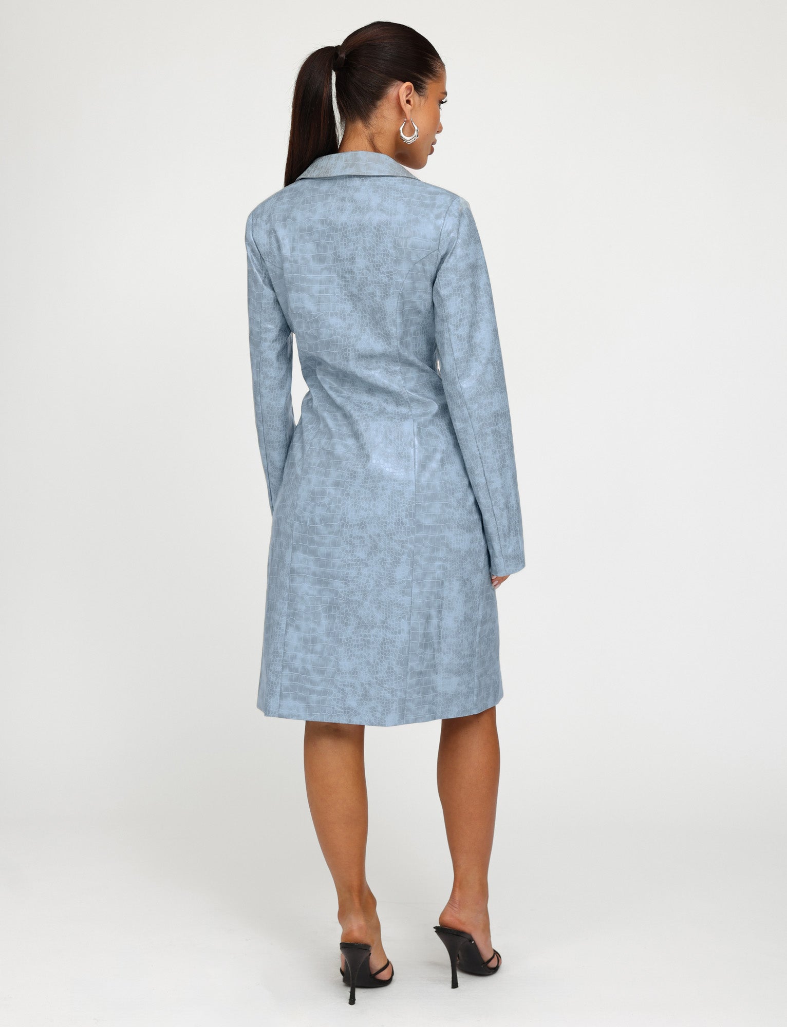 A woman with a ponytail is standing with her back to the camera, showcasing a TIGERMIST EMERSYN TRENCH COAT in dusty blue, paired elegantly with black high-heeled sandals against a plain white background.