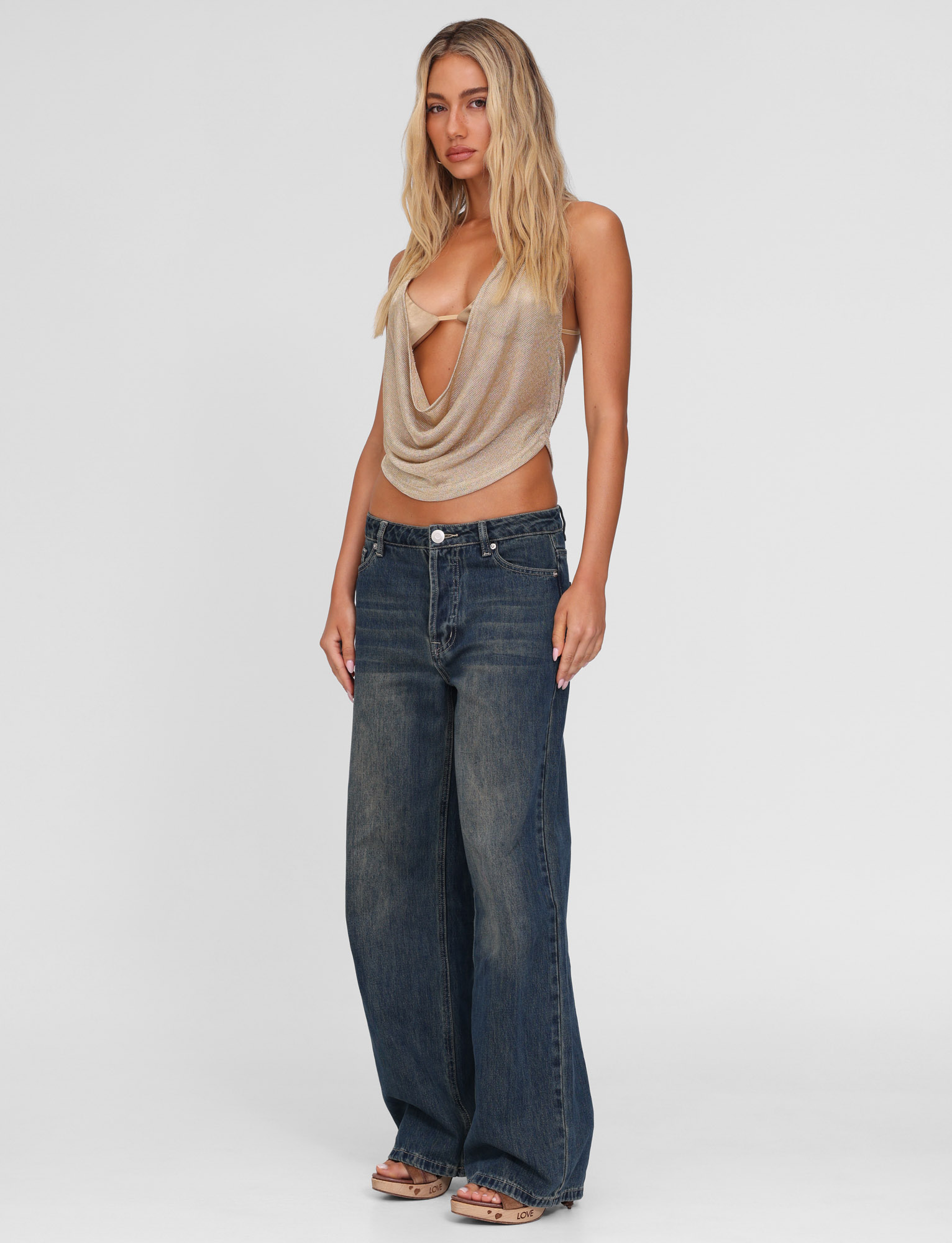 A woman with long, wavy blonde hair wears the TIGERMIST FELIX JEAN - BLUE : MID WASH, paired with a draped, sleeveless beige top. She stands facing forward with a neutral expression against a plain white background.