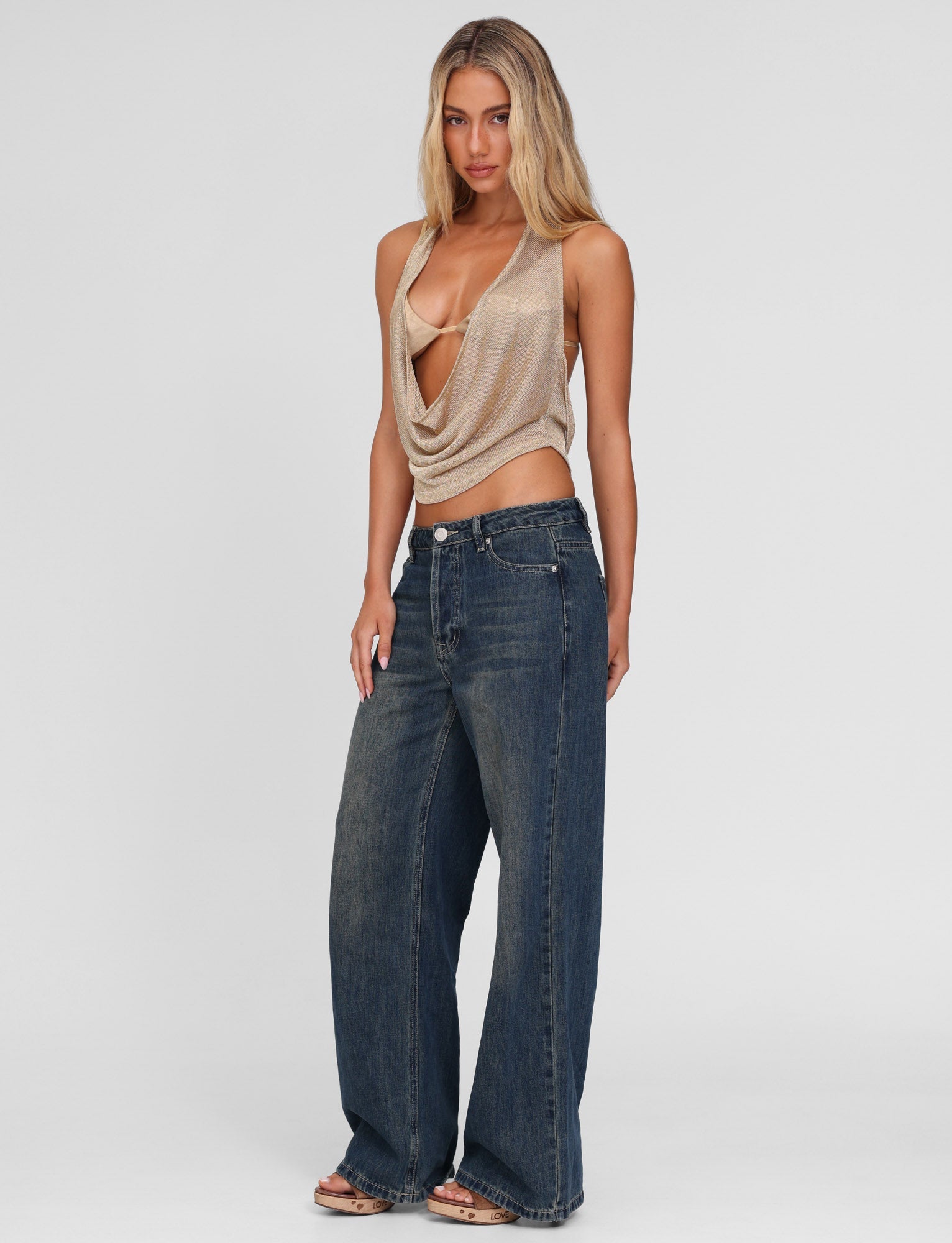 A woman with long blonde hair stands against a plain background wearing TIGERMIST’s FELIX JEAN - BLUE : MID WASH, styled high-waisted and wide-leg, paired with a draped beige halter top and sandals.