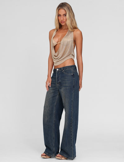 A woman with long blonde hair stands against a plain background wearing TIGERMIST’s FELIX JEAN - BLUE : MID WASH, styled high-waisted and wide-leg, paired with a draped beige halter top and sandals.