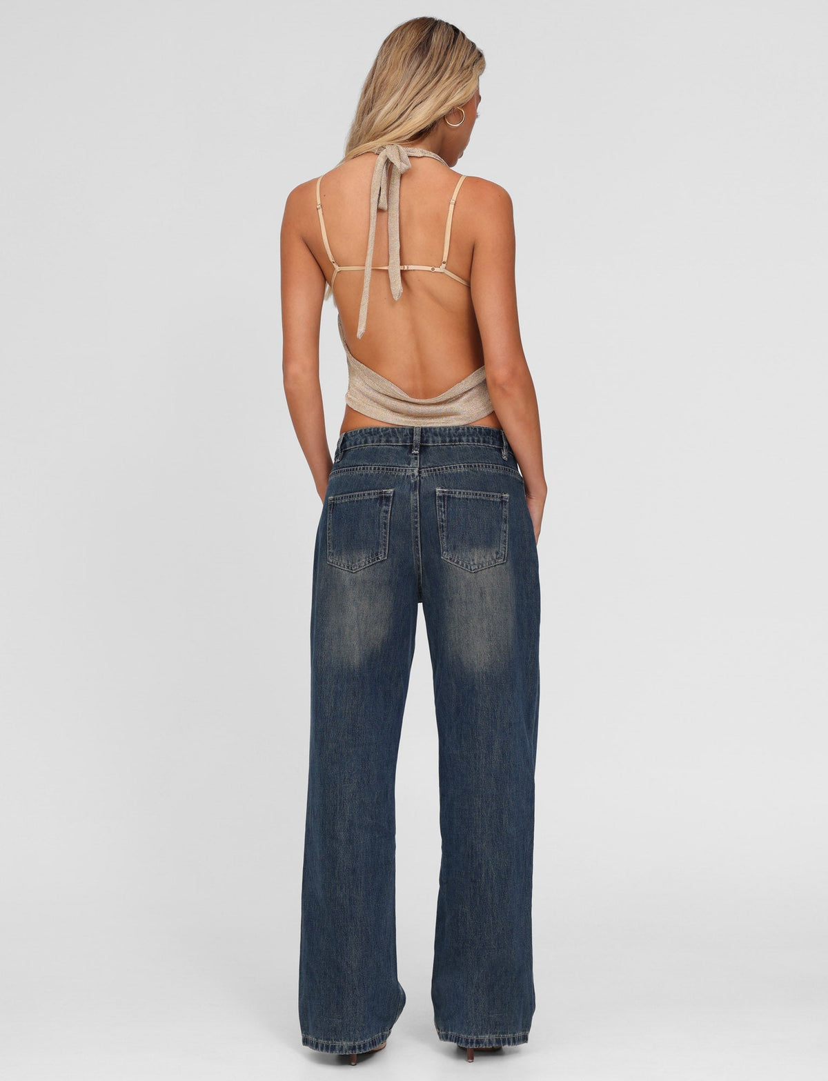 A woman with long blonde hair stands facing away, hands in pockets, wearing TIGERMIST FELIX JEAN - BLUE : MID WASH and a beige backless halter top against a plain white background.