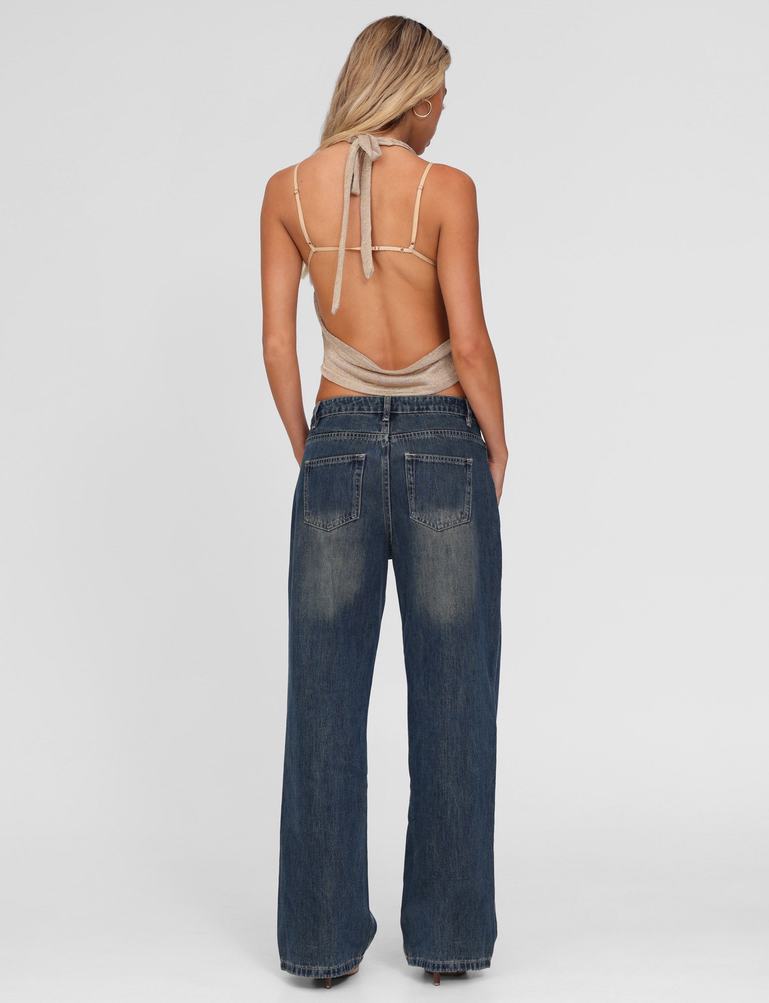 A woman with long blonde hair stands facing away, hands in pockets, wearing TIGERMIST FELIX JEAN - BLUE : MID WASH and a beige backless halter top against a plain white background.