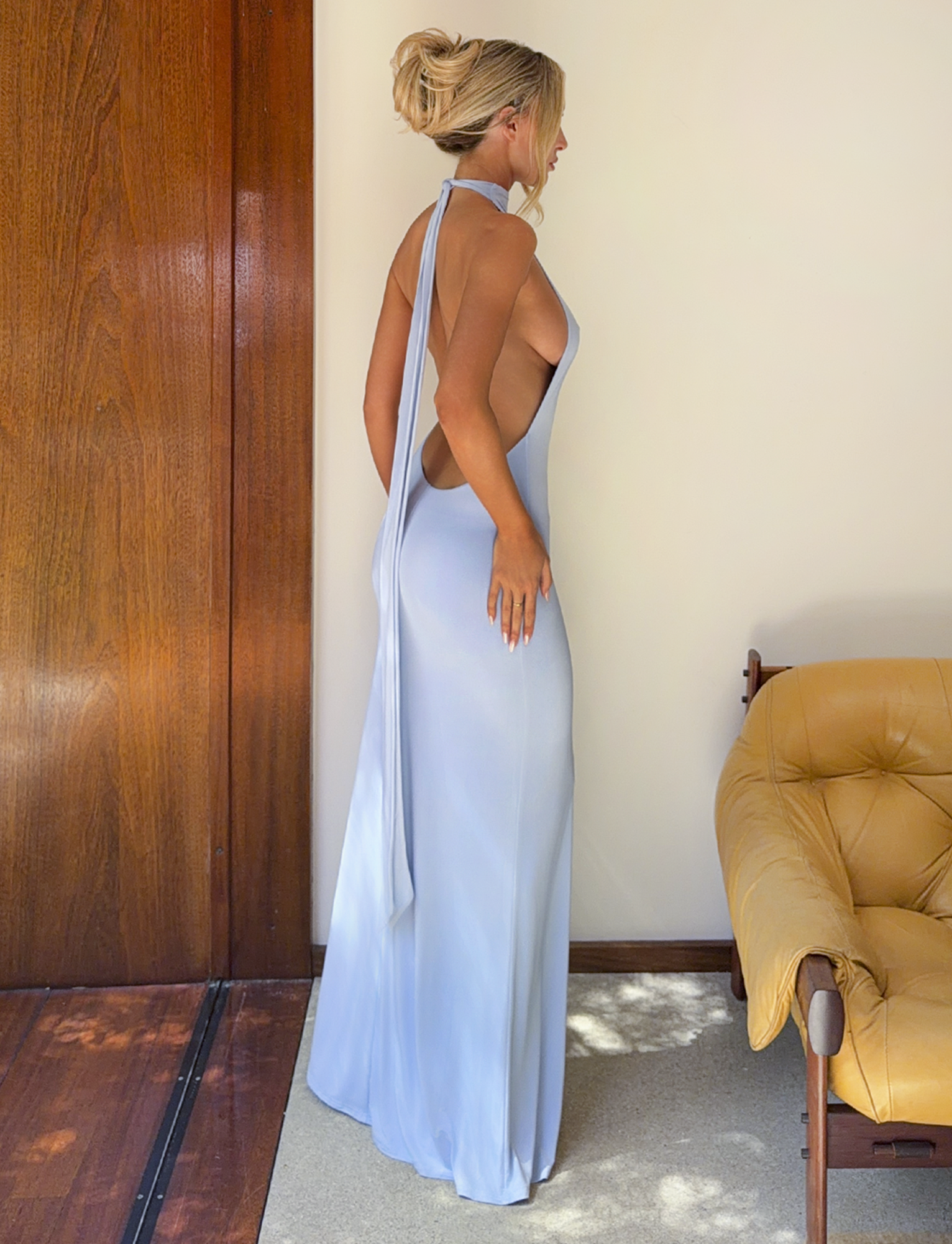 A woman with blonde hair in a bun poses indoors, wearing the TIGERMIST MADDY MAXI DRESS in baby blue. She stands sideways beside a wooden wall and mustard yellow chair as sunlight highlights the dress’s elegant open back.
