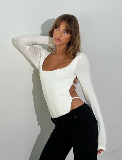 A woman with light brown hair poses against a white wall. She wears the TIGERMIST MINERVA LONG SLEEVE BODYSUIT in white, along with black high-waisted pants from Tiger Mist. One arm is raised behind her head, and she gazes to the side with a relaxed expression.