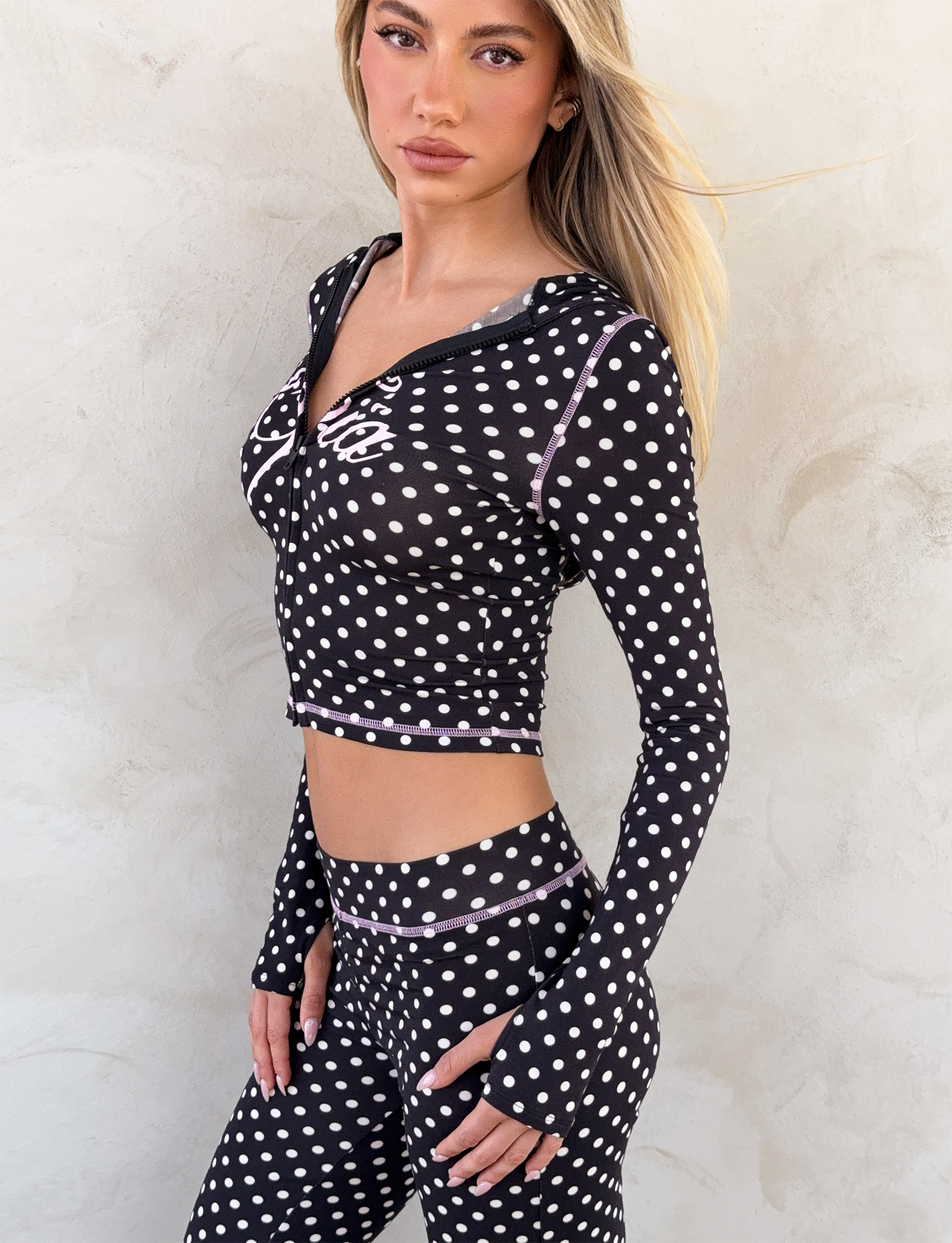 A woman in the I.AM.GIA BLARE HOODIE - BLACK POLKA DOT and matching high-waisted leggings with white polka dots poses against a light textured wall.