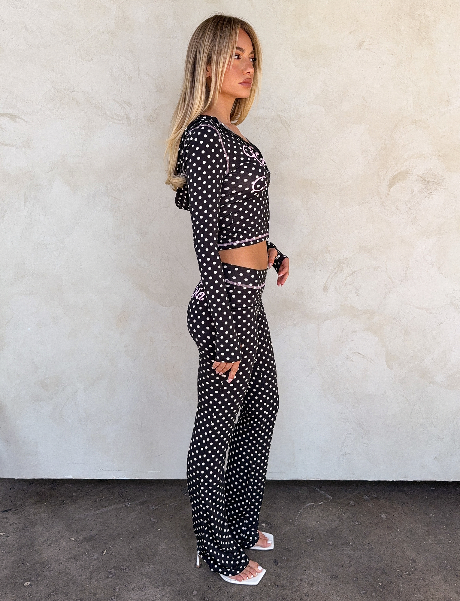 A woman stands sideways against a light textured wall, wearing I.AM.GIA’s BLARE TRACKPANT in black polka dot and a matching black crop top, paired with white open-toe heels.
