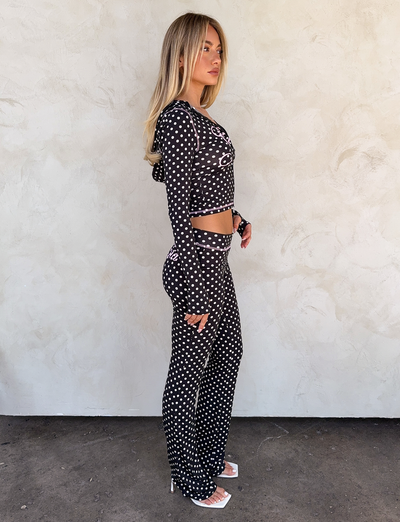 A woman stands sideways against a light textured wall, wearing I.AM.GIA’s BLARE TRACKPANT in black polka dot and a matching black crop top, paired with white open-toe heels.