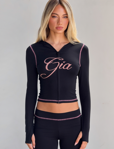 A woman with long blonde hair models the Tiger Mist CL - BLARE HOODIE, a fitted black zip-up top with pink trim and "Gia" on the chest, paired with matching black pants, against a plain light gray background.