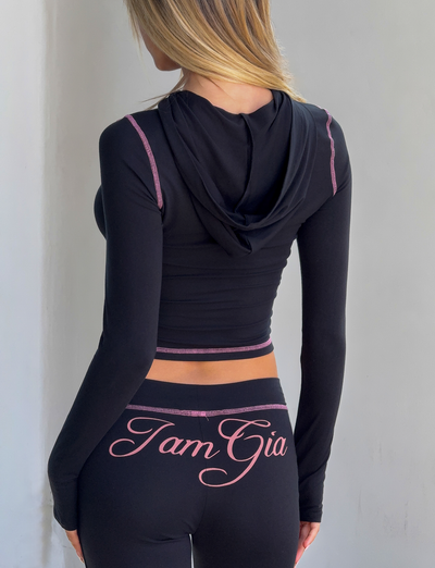 A blonde woman is seen from behind wearing the I.AM.GIA BLARE HOODIE in black and matching leggings with "TamGia" printed in cursive on the waistband.
