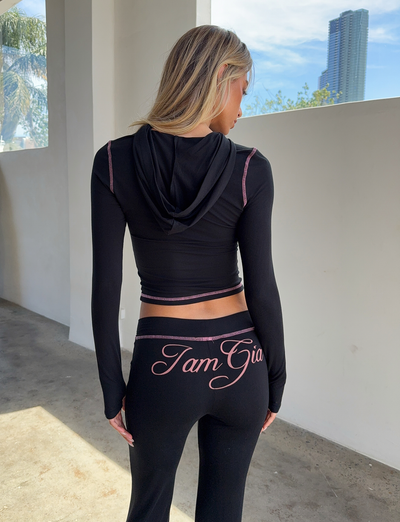 A woman stands indoors with her back to the camera, wearing the I.AM.GIA BLARE HOODIE in black and matching pants. Sunlight streams through windows with city buildings visible outside.