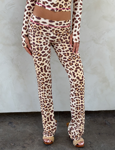 A person stands against a light textured wall, wearing I.AM.GIA's BLARE TRACKPANT in NEUTRAL : LEOPARD : WILD CARD, paired with a matching long-sleeve crop top. Their hands rest near their thighs.