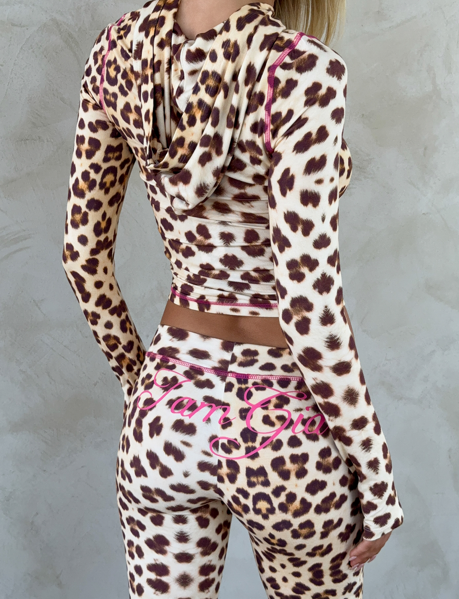A woman faces away from the camera wearing I.AM.GIA's BLARE TRACKPANT in Neutral Leopard Wild Card, featuring pink script on the back, paired with a matching hooded top against a light textured background.