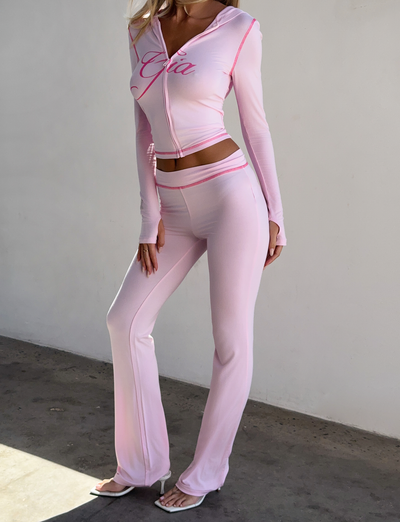 A woman indoors wears I.AM.GIA's BLARE TRACKPANT in pastel pink, paired with a matching hooded zip-up crop top and white heeled sandals. Her face is not visible.