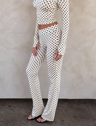 A person, face unseen, stands against a light wall wearing a fitted long-sleeve top and the I.AM.GIA BLARE TRACKPANT in cream polka dot.