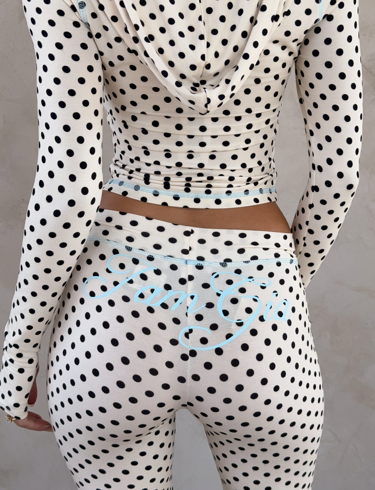 A person faces away from the camera wearing the I.AM.GIA BLARE TRACKPANT in cream polka dot, a fitted, white hooded outfit with polka dots. Light blue cursive "I am Gia" text appears on the back of the trackpant.