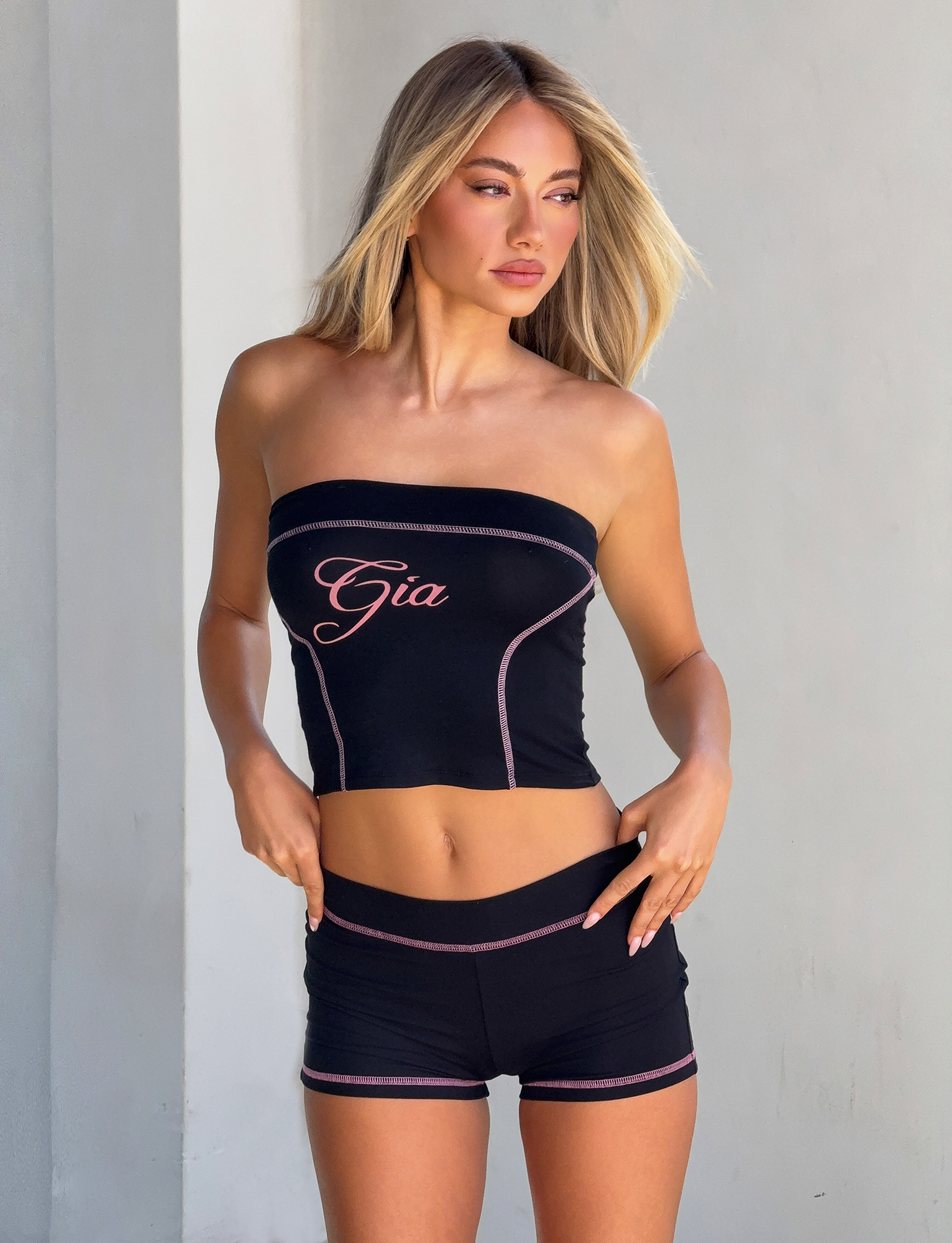A woman with long blonde hair models the Tiger Mist CL - BLARE SHORT and a black strapless crop top with "Gia" lettering, standing against a plain gray background.