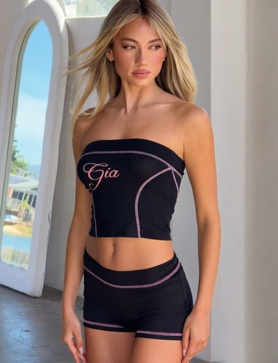 A woman with long blonde hair wears a black tube top with "Gia" in pink script and I.AM.GIA's BLARE SHORT - BLACK, featuring pink trim, while standing indoors by a large arched window as sunlight streams in.