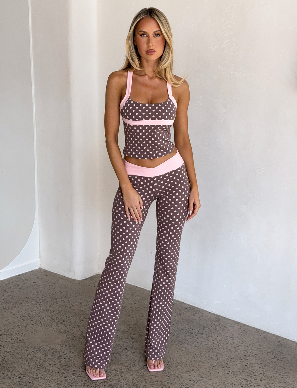 A woman poses indoors against a light wall, wearing the I.AM.GIA BLARE HALTER STRAP TOP in pink chocolate with matching flared pants featuring white polka dots and pink trim from the Blare collection.