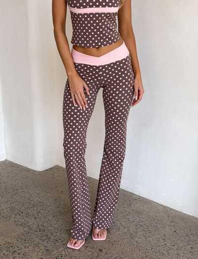 A woman, visible from the neck down, stands indoors wearing a brown sleeveless polka dot crop top and I.AM.GIA BLARE CROSSOVER PANT - PINK CHOCOLATE with pink sandals. Her hands rest by her sides.