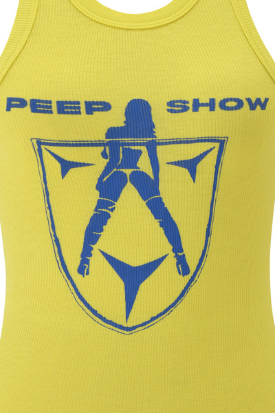 The I.AM.GIA PENELOPE TANK - BLUE PEEP SHOW features a ribbed yellow design with a blue shield graphic of a posing woman and bold "PEEP SHOW" text, channeling the edgy style of I.AM.GIA.