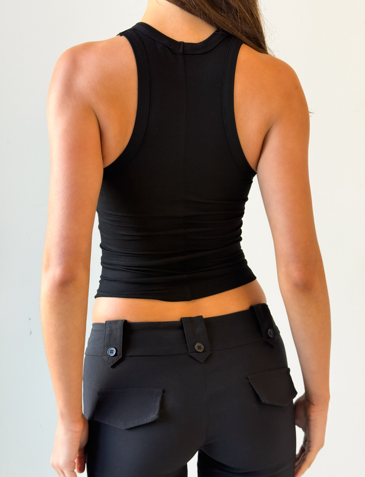 A woman is shown from the back, wearing the I.AM.GIA PENELOPE TANK - GIA LOGO in sleek black and black pants with buttoned back pockets against a plain, light-colored background.