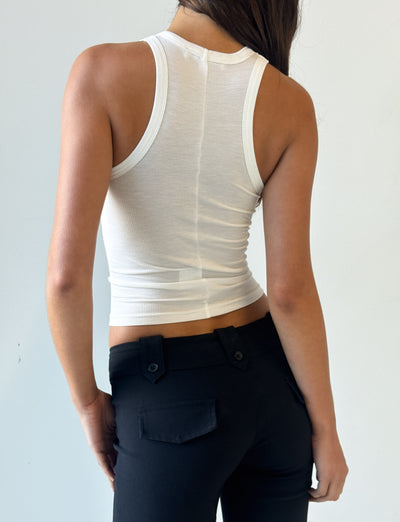 A person stands facing away, wearing the I.AM.GIA PENELOPE TANK - PINK AIRBRUSH with ribbed fabrication and black pants with buttoned pockets, set against a plain light background.
