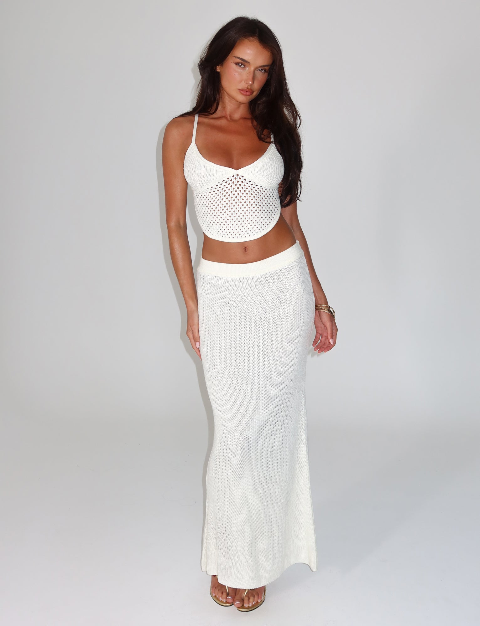 A woman stands barefoot against a plain background, wearing the TIGERMIST white crochet crop top paired with the RAMSEY SKIRT in white. Her long dark hair cascades freely as she gracefully places one hand on her waist.