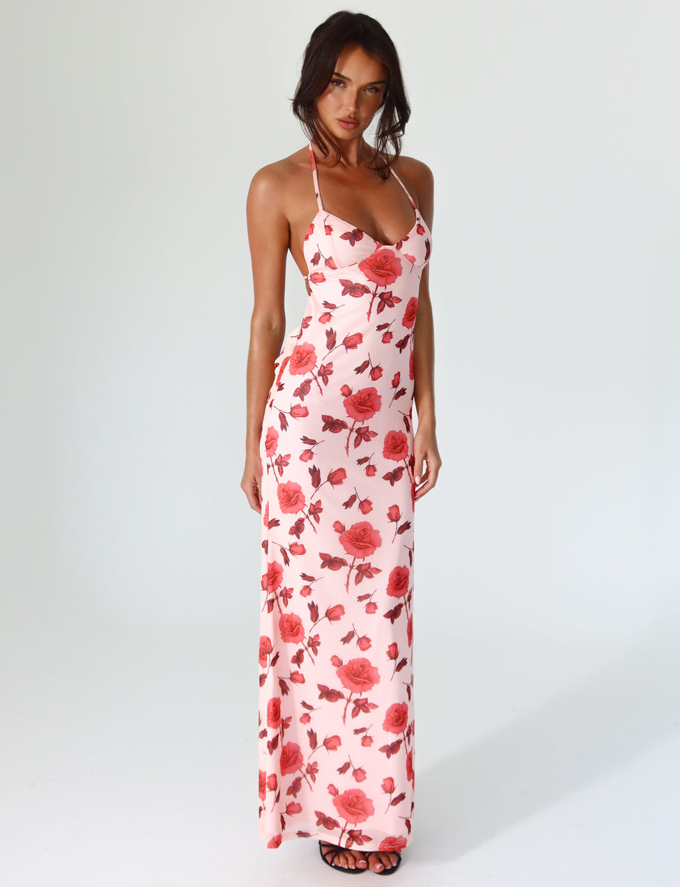 A person is wearing the BRYSTOL DRESS - RED : FLORAL by TIGERMIST, a full-length, sleeveless maxi dress featuring a halter neckline and a striking red rose pattern. They are standing against a plain background, paired with black sandals. Their long dark hair flows down as they look directly at the camera.