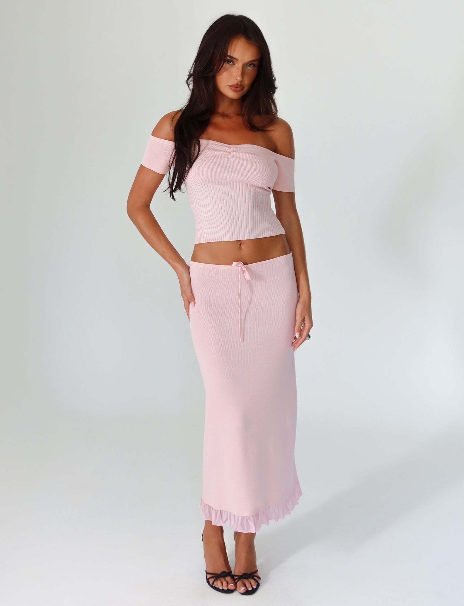 A woman is posing in the Lilyanna Skirt - Baby Pink from Tigermist, featuring a matching light pink off-the-shoulder top with a relaxed fit and ruffled hem midi skirt. She pairs the outfit with black open-toed heels and has long dark hair, maintaining a neutral expression against a plain white background.