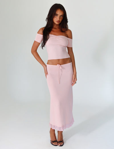 A woman is posing in the Lilyanna Skirt - Baby Pink from Tigermist, featuring a matching light pink off-the-shoulder top with a relaxed fit and ruffled hem midi skirt. She pairs the outfit with black open-toed heels and has long dark hair, maintaining a neutral expression against a plain white background.