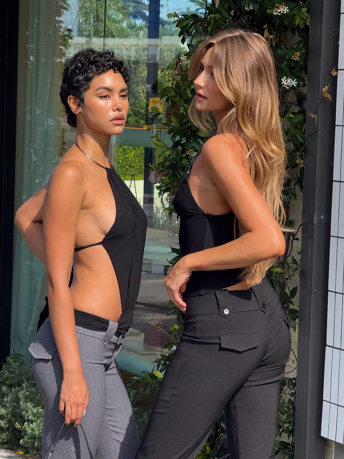 Two women pose outdoors in stylish black TIGERMIST looks—one wears the FENELLA TOP - BLACK, the other fitted pants. With greenery and a building behind them, they stand confidently; one has short curly hair, the other long and wavy.
