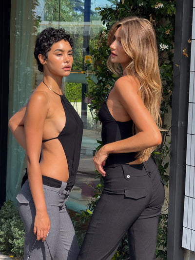 Two women pose outdoors in stylish black TIGERMIST looks—one wears the FENELLA TOP - BLACK, the other fitted pants. With greenery and a building behind them, they stand confidently; one has short curly hair, the other long and wavy.