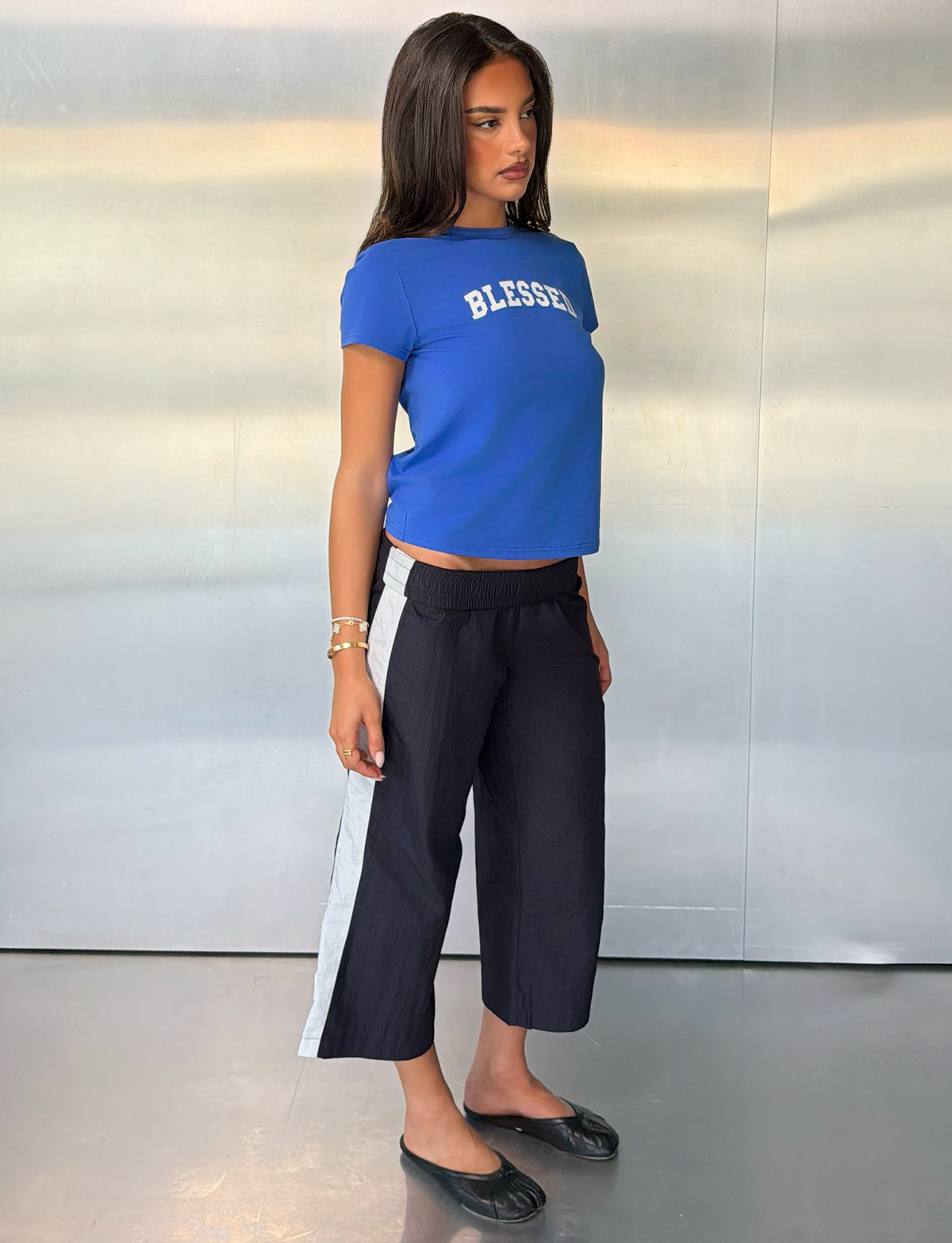 A woman stands in front of a metallic wall, wearing the TIGERMIST BLESSED BABY TEE - BLUE, which features a vintage relaxed fit. She pairs it with black and white side-striped cropped pants, black flats, and bracelets. With a neutral expression, she gazes slightly to the side.