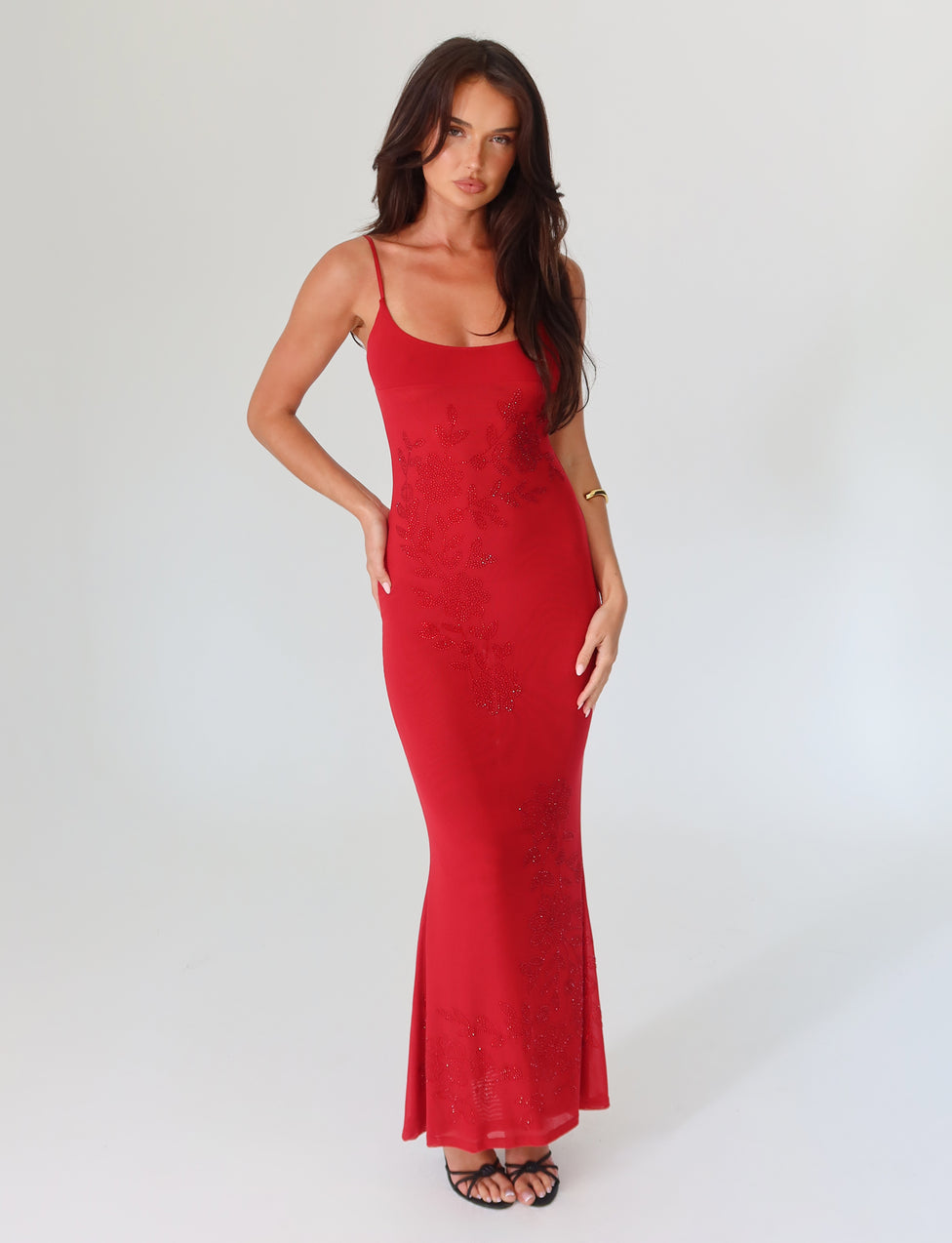 A woman stands confidently in a sleeveless, form-fitting Cherry Red Octavia Maxi Dress by Tigermist with subtle floral embellishments. She wears black high heels and has long, dark hair, posing against a plain light background.