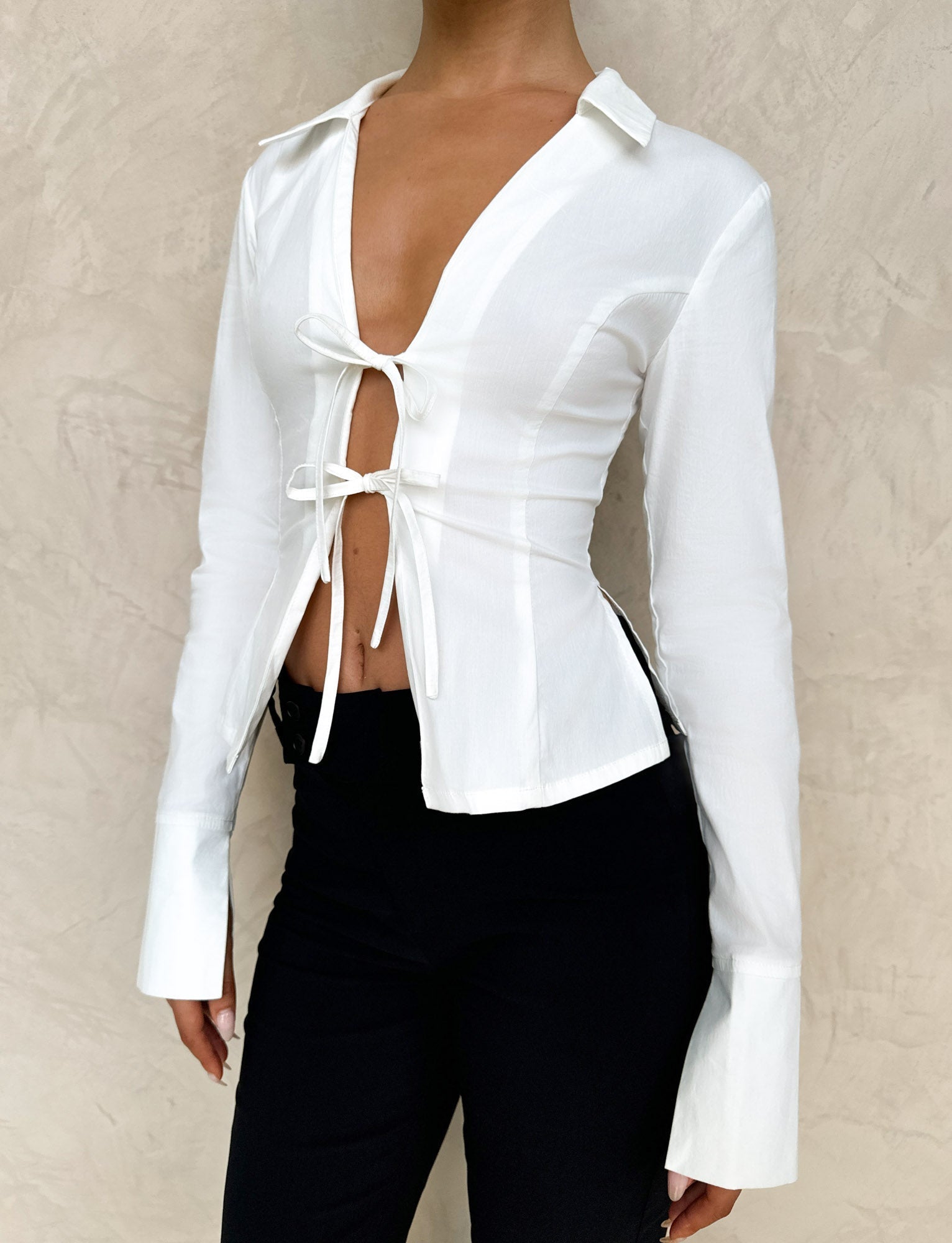 Someone is wearing the TIGERMIST BRIELLE SHIRT - WHITE : IVORY, featuring front ties and a deep neckline, paired with black pants against a textured light beige wall background.