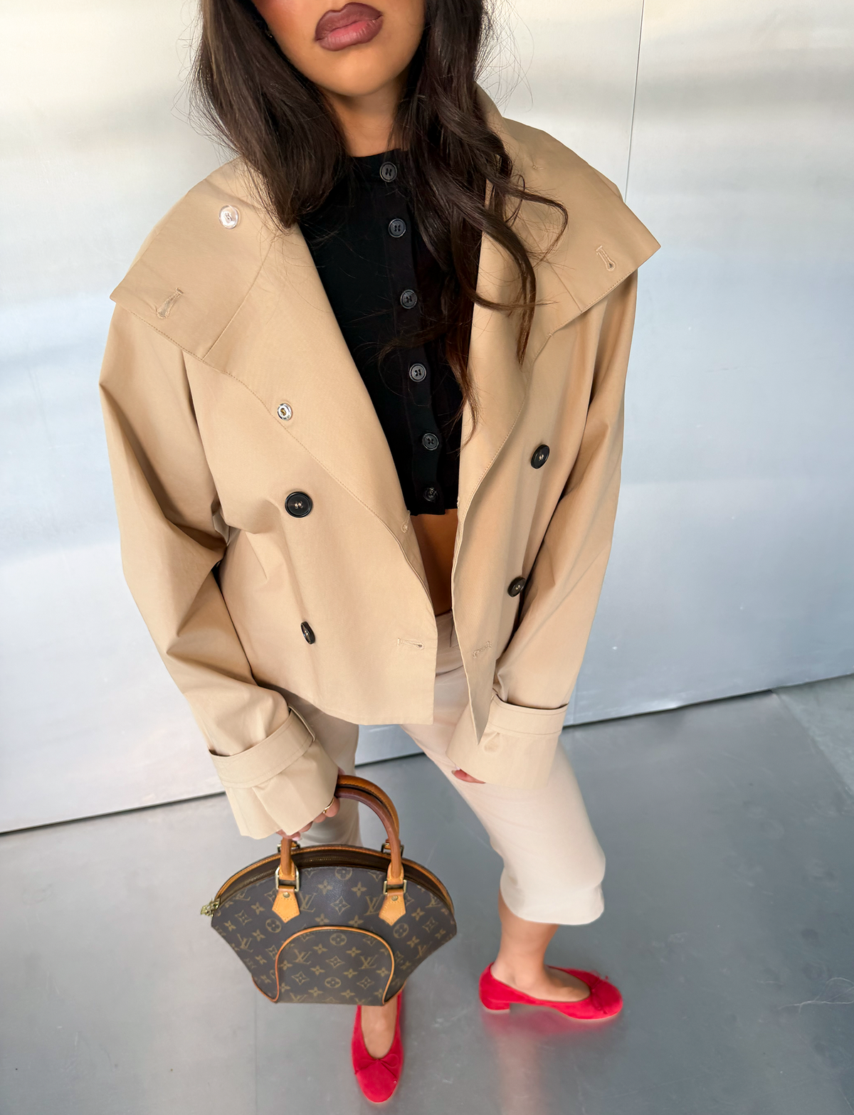 A woman wears the TIGERMIST HAYLEY Cropped Trench Jacket in khaki, styled with cropped pants and a black top. She holds a brown designer handbag and stands on a reflective floor, her loose hair partially obscuring her face, while finishing the look with bright red flats.