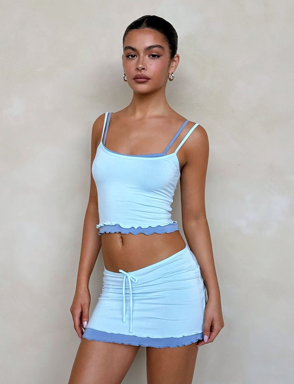A woman stands against a plain taupe background wearing the ADAIR SKIRT - BLUE by TIGERMIST, paired with a light blue camisole featuring double shoulder straps. Both the skirt and camisole have ruffled light blue hems. She accessorizes with gold hoop earrings and has her hair pulled back.
