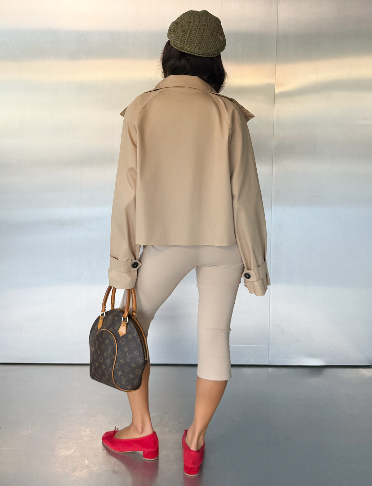 A woman seen from behind wears the TIGERMIST HAYLEY CROPPED TRENCH JACKET in khaki, featuring a boxy fit. She pairs it with light capri pants, a green cap, red loafers, and carries a brown handbag while standing in front of a metallic background.