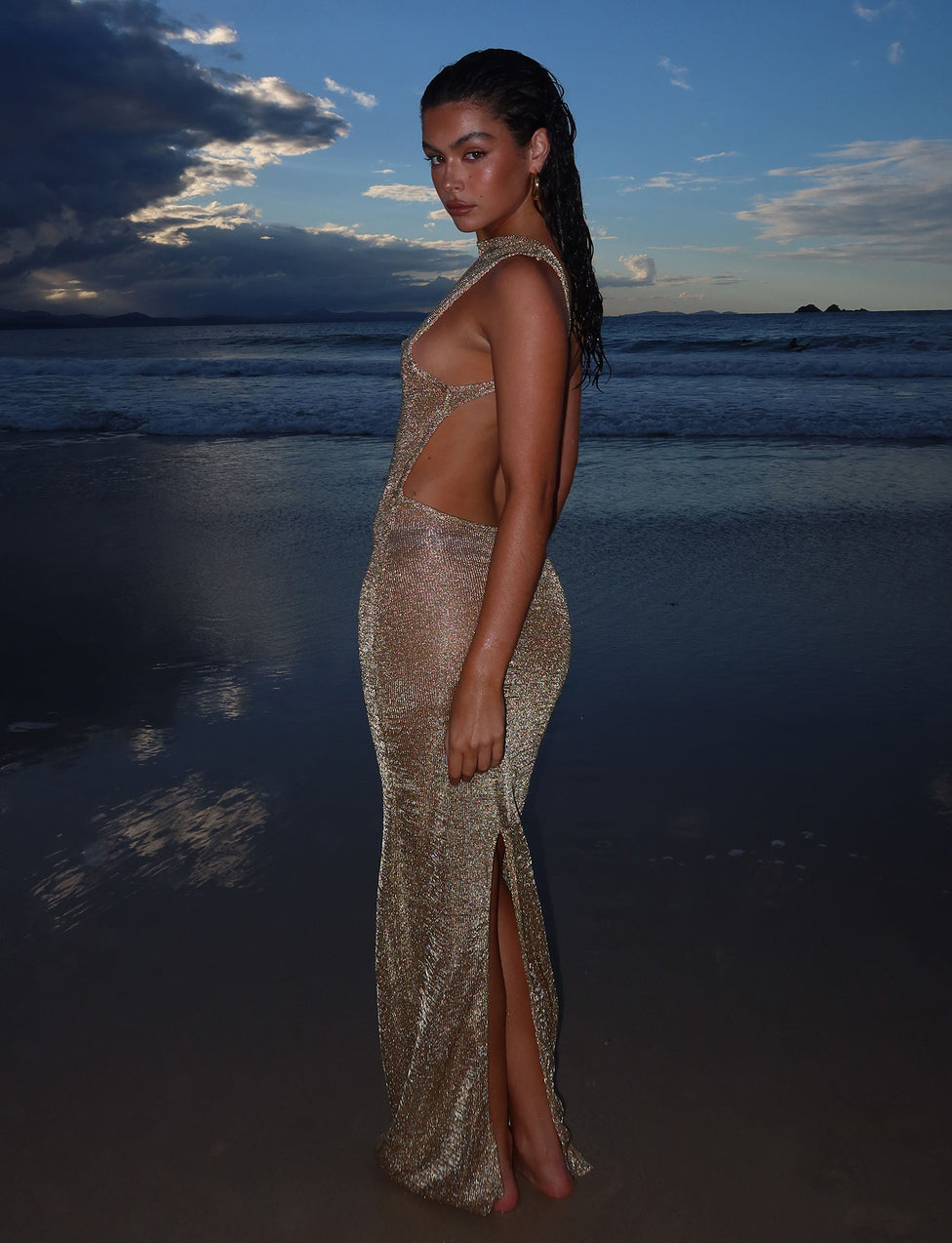 A woman stands on a beach at dusk, wearing the shimmering, semi-sheer ZAELA DRESS - GOLD from TIGERMIST, with cutouts and a thigh-high slit. The waves gently lap at the shore behind her, and a dramatic, cloud-filled sky meets the horizon. Her hair appears wet as she poses confidently.
