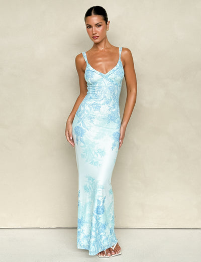 A woman poses in front of a neutral backdrop, wearing the TIGERMIST GUILIANNA FRILL MAXI DRESS - BLUE : FLORAL : TAPESTRY, a form-fitting and sleeveless piece crafted from high stretch fabric. Her hair is pulled back and she pairs the elegant V-neck design with white sandals, highlighting its adjustable frill shoulder straps that add an extra touch of charm.