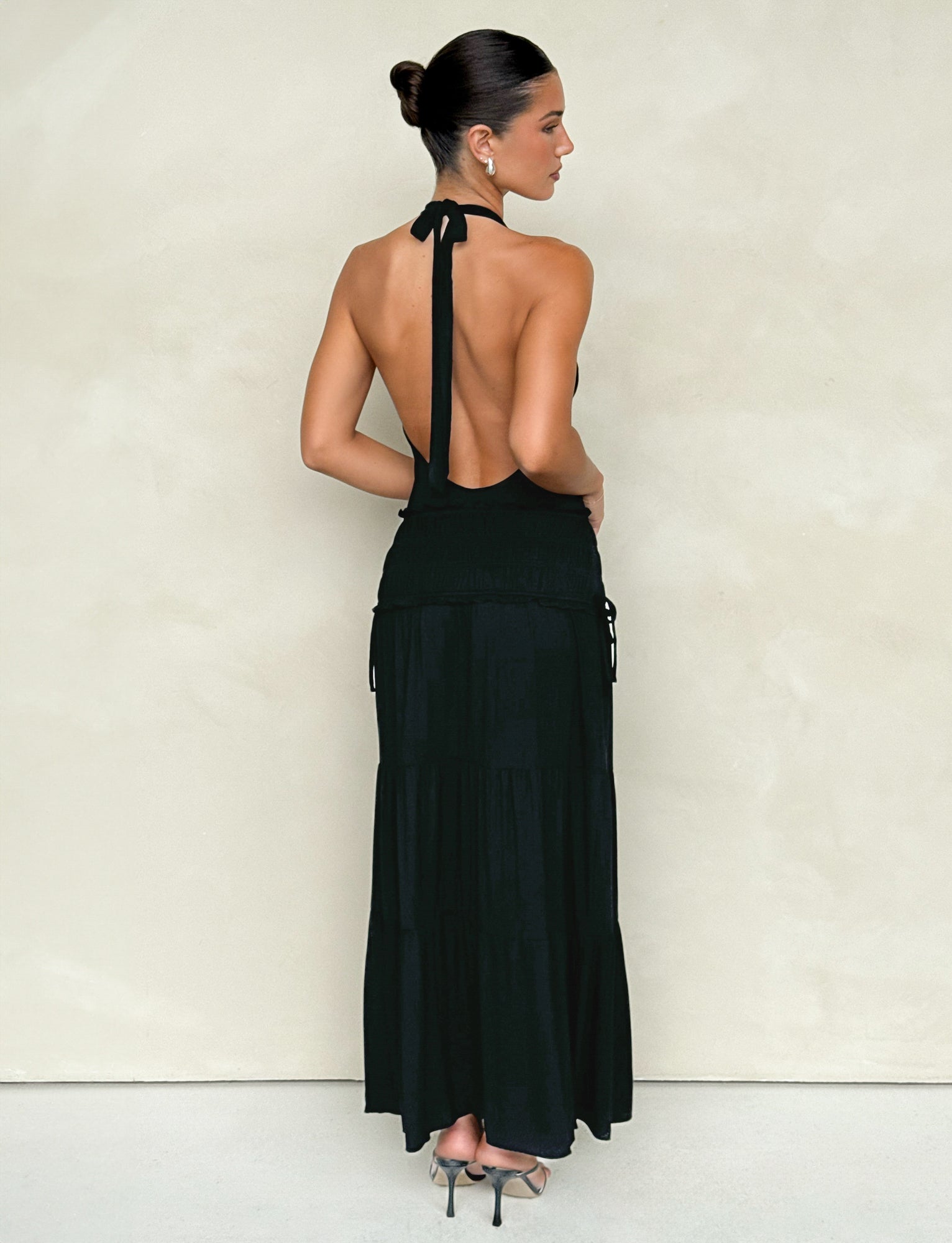 A woman with dark hair styled in a bun is wearing the DENALI HALTER MAXI DRESS by TIGERMIST, paired with high heels. She stands against a simple, light-colored wall, showcasing the elegant low back of the sleek black dress as she faces away from the camera.