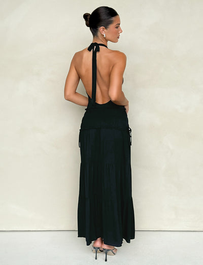 A woman with dark hair styled in a bun is wearing the DENALI HALTER MAXI DRESS by TIGERMIST, paired with high heels. She stands against a simple, light-colored wall, showcasing the elegant low back of the sleek black dress as she faces away from the camera.