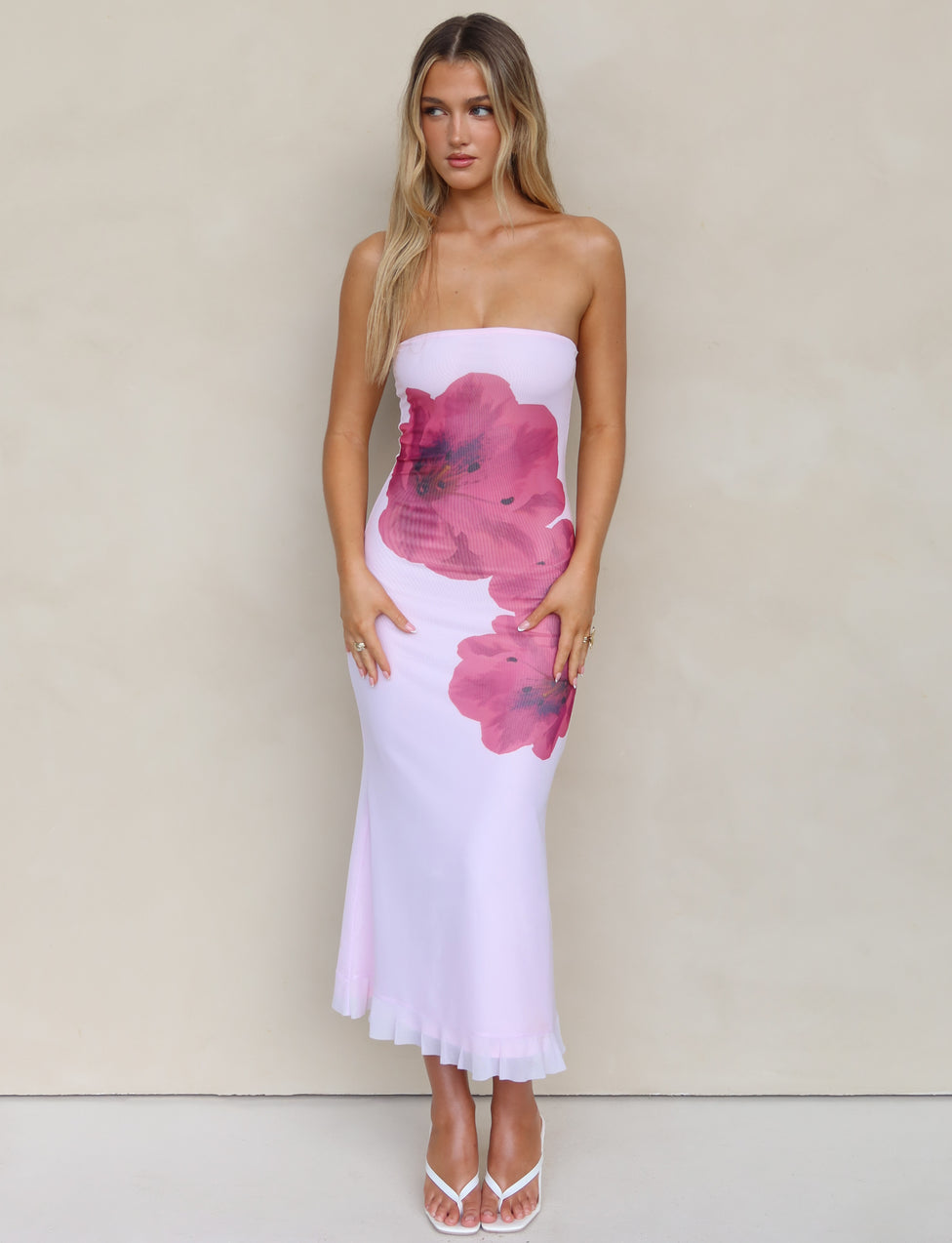 A woman with long blonde hair is wearing the LILLIAN DRESS - PINK by TIGERMIST, a strapless white dress adorned with large pink floral designs. She stands against a plain, beige background, complemented by her white, open-toed high heels. Her arms hang by her side as she gazes slightly downward, showcasing the elegant midi length of her outfit.