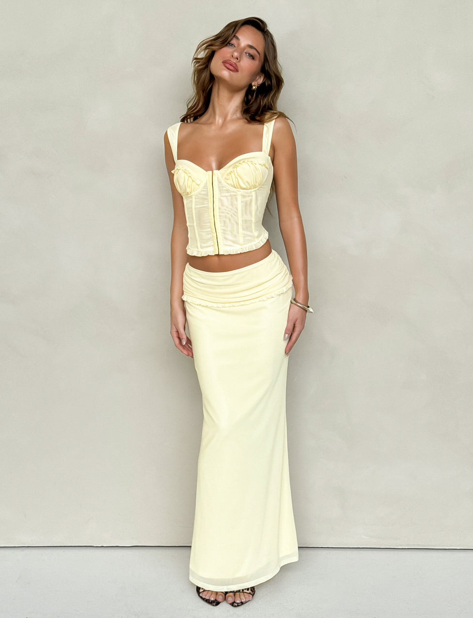 A woman is posing against a plain backdrop, wearing a fitted yellow corset top and the TIGERMIST GINNY MAXI SKIRT - LEMON, which features a wide waistband. She stands with her head tilted and her hair flowing freely.